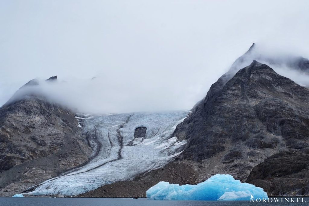 dicke wolken über rauhen bergen und einem gletscher an der küste mit einem blauen eisberg